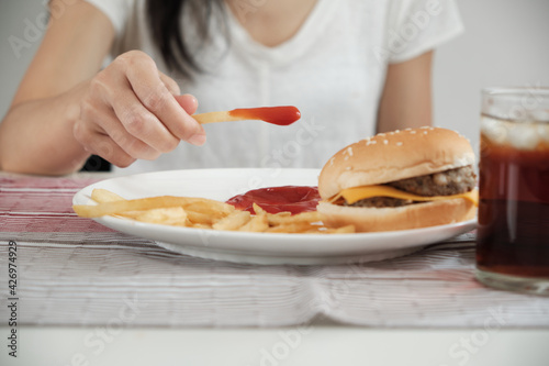 Close-up shot: A piece of French fire in a woman's hand-dipped in ketchup eaten along with other unhealthy junk food like a hamburger and cola in a white dish which is placed on red tablecloth.