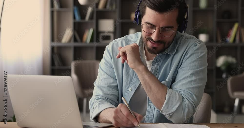 Concentrated smart young male worker employee entrepreneur in eyeglasses wearing wireless headphones, holding negotiations meeting online with client or colleagues, using computer zoom video call app.