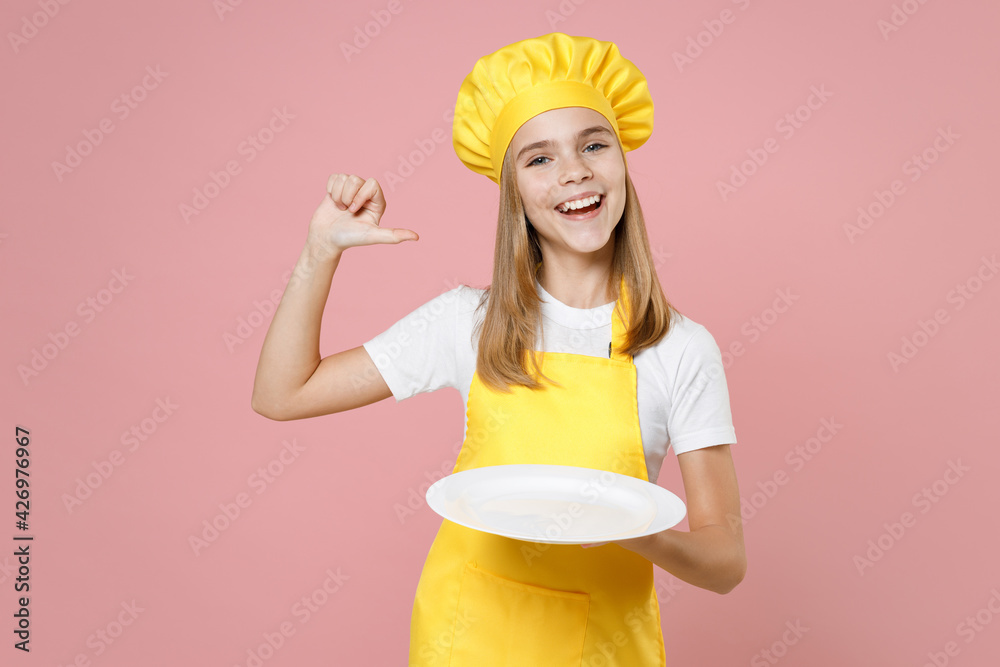 Teen girl chef cook confectioner housewife baker in yellow apron white t-shirt cap hold empty blank plate, place for food point thumb finger on herself isolated on pink background Food cake concept. Teen girl chef cook confectioner housewife baker in yellow apron white t-shirt cap hold empty blank plate, place for food point thumb finger on herself isolated on pink background Food cake concept.