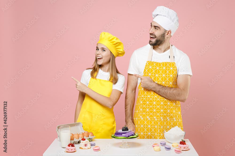 Teen fun girl father chef cook confectioner baker wear yellow apron toque cap at table point finger aside on workspace area isolated on pink background. Mousse cake food workshop master class process. Teen fun girl father chef cook confectioner baker wear yellow apron toque cap at table point finger aside on workspace area isolated on pink background. Mousse cake food workshop master class process.