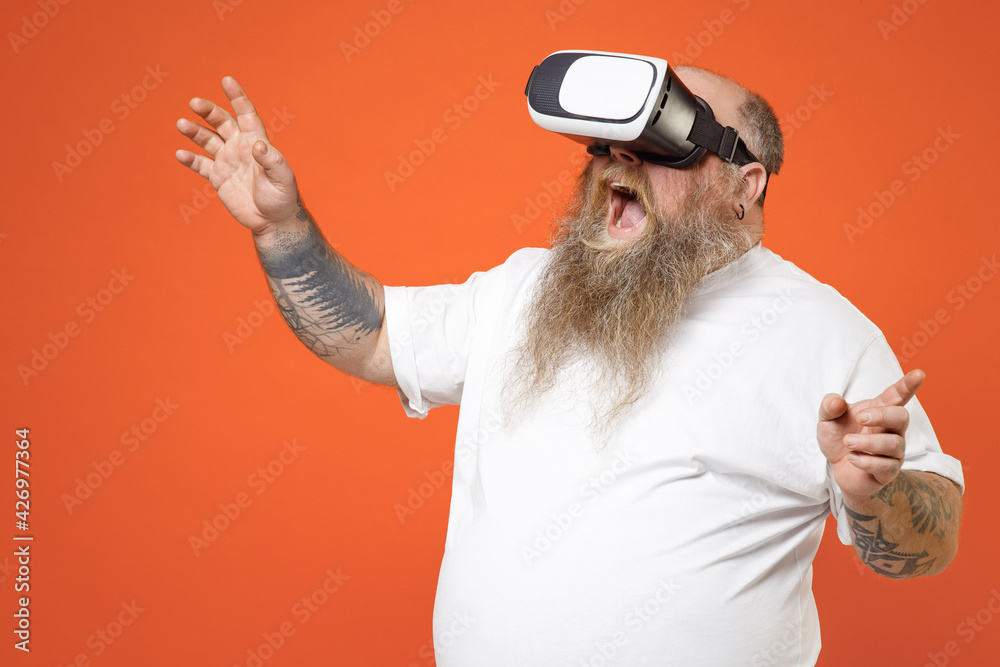 Naklejka premium Fat fun happy pudge obese chubby overweight tattooed bearded man 30s has big belly wear white t-shirt watching in vr headset pc gadget stretching hands isolated on orange background studio portrait