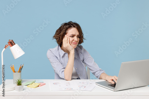 Young puzzled tired frustrated secretary employee business woman in casual shirt sit work at white office desk use pc laptop computer internet prop up face isolated on pastel blue background studio.