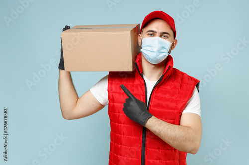 Delivery guy employee man in red cap white t-shirt vest uniform sterile face mask gloves work courier service on lockdown covid19 flu virus hold empty cardboard box isolated on pastel blue background