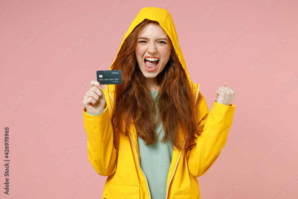 Redhead young woman 20s in yellow waterproof raincoat hood outerwear do winner gesture clench fist hold credit bank card isolated on pastel pink background studio. Lifestyle wet fall season concept Redhead young woman 20s in yellow waterproof raincoat hood outerwear do winner gesture clench fist hold credit bank card isolated on pastel pink background studio. Lifestyle wet fall season concept