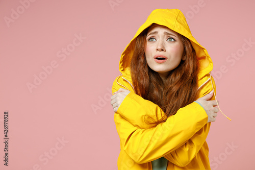 Redhead young frozen woman in yellow waterproof raincoat hood hug herself look aside outerwear isolated on pastel pink color background studio Outdoors lifestyle wet fall cold weather season concept