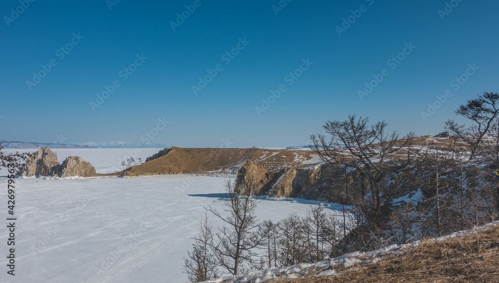 The surface of the frozen lake with steep banks is covered with snow. A picturesque two-headed rock rises above the ice. Clear blue sky. On the ground are bare trees. Baikal The surface of the frozen lake with steep banks is covered with snow. A picturesque two-headed rock rises above the ice. Clear blue sky. On the ground are bare trees. Baikal