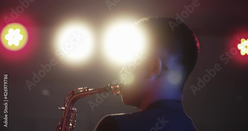 A young jazz musician wearing the black glasses , play saxophones on a stage in the club at night, lit up in beautiful red, yellow and blue lights. photo