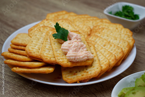 Close-up of cheese crackers are spread out on a plate. On one cracker, spread and parsley.
