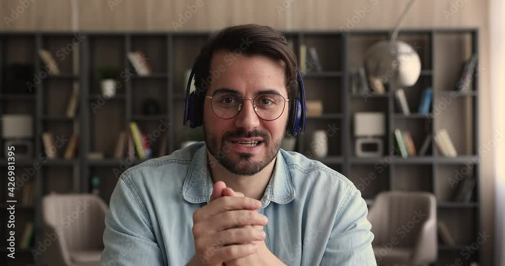 Smiling young handsome caucasian man in eyeglasses wearing modern wireless headphones, looking at camera, holding zoom video call conversation, talking speaking negotiating project, gadget screen view