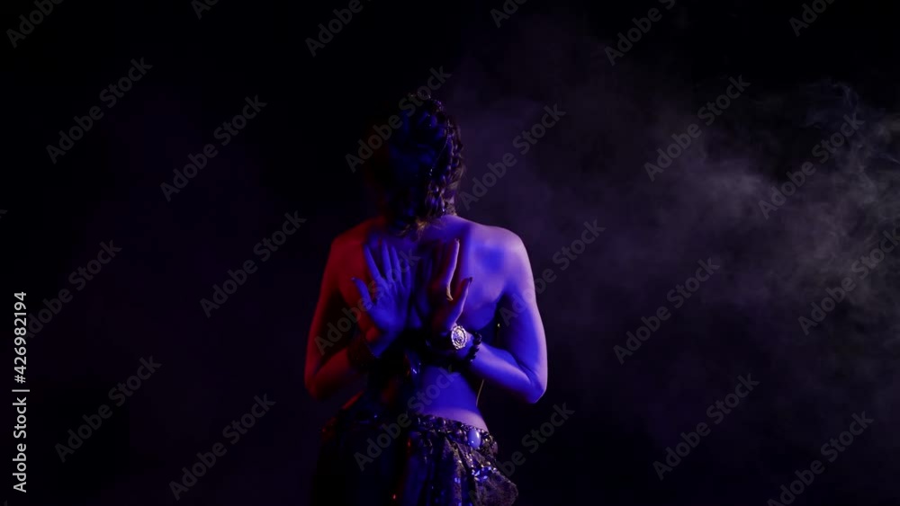 A woman, seen from the back, dancing an oriental dance in a smoky studio on a black background, makes beautiful movements with her hands, she is dressed in a chic suit and illuminated by neon light. A woman, seen from the back, dancing an oriental dance in a smoky studio on a black background, makes beautiful movements with her hands, she is dressed in a chic suit and illuminated by neon light.