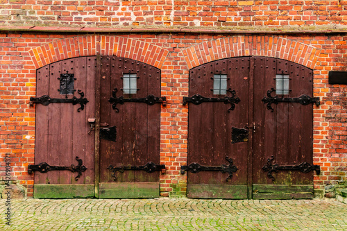 Close up image of a couple of wooden doors of a historical red brick building, with metal supports and locks, with moss on the wall and on the ground.