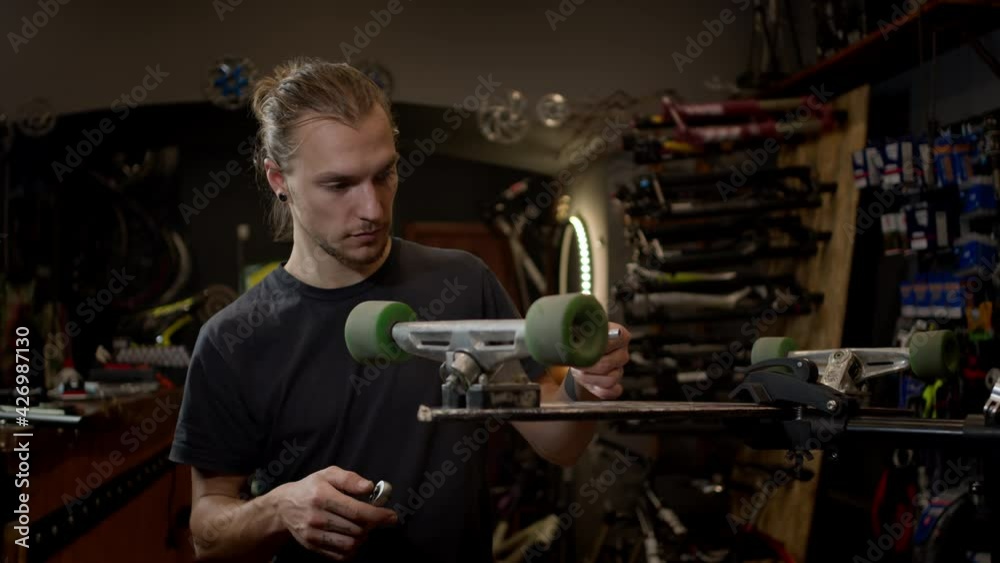 Young Man In The Workshop Attaches Wheel To Skateboard. Many Tools And Parts In The Workshop. Handsome Guy With Beard Works Carefully. Young Man In The Workshop Attaches Wheel To Skateboard. Many Tools And Parts In The Workshop. Handsome Guy With Beard Works Carefully.