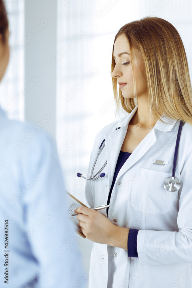 Young woman-doctor and her patient are discussing patient's current health examination, while standing together in a hospital office. Female physician is writing some marks, using a clipboard. Perfect Young woman-doctor and her patient are discussing patient's current health examination, while standing together in a hospital office. Female physician is writing some marks, using a clipboard. Perfect