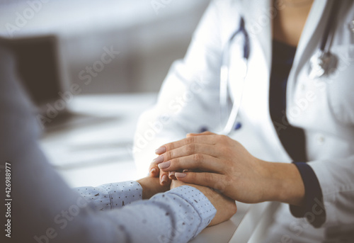 Unknown woman-doctor is holding her patient s hands  discussing current health examination  while they are sitting at the desk in the cabinet in a clinic. Physician with a stethscope at work  close-up