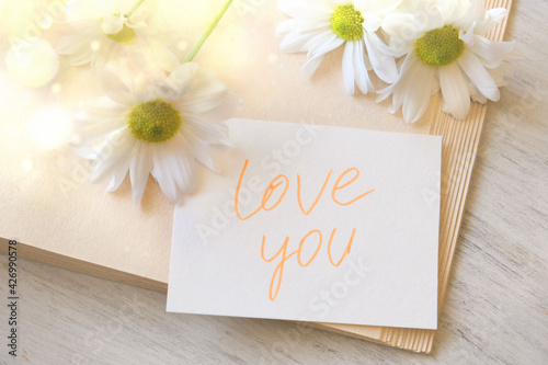 Handwritten note Love You and summer flowers. Sun shine and glare on bouquet of chamomiles. Soft focus