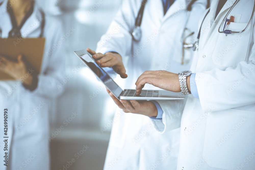 Group of unknown doctors, men and a woman, with stethoscopes, discuss medical exam resoults, using a laptop, while standing at hospital office. Physicians using a laptop for checking up medication Group of unknown doctors, men and a woman, with stethoscopes, discuss medical exam resoults, using a laptop, while standing at hospital office. Physicians using a laptop for checking up medication