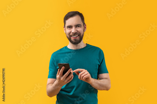 Studio portrait of young smiling bearded man holding smartphone in hands and making bets online at bookmaker's website using mobile application. Isolated over yellow background