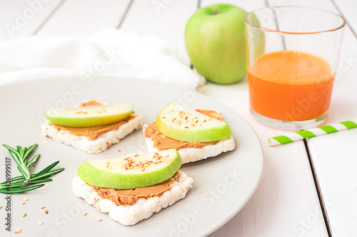 Healthy breakfast with peanut butter and apple sandwiches on rice cakes, carrot juice and apple on white wooden background