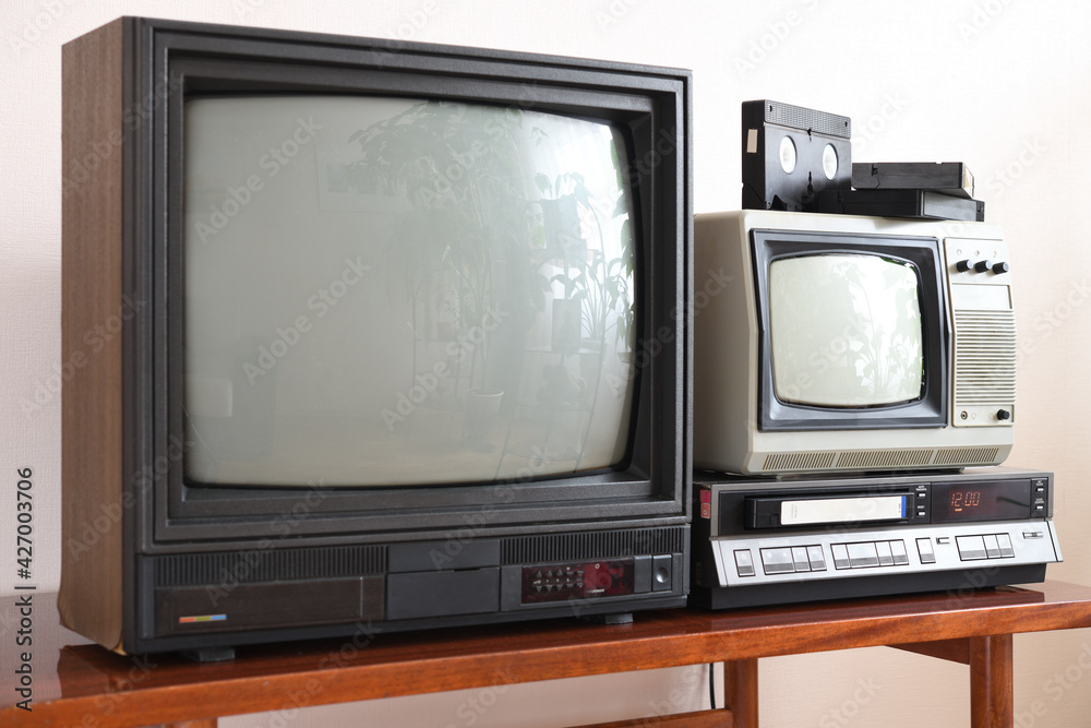 Two antiquated vintage TVs with VCRs sit on a vintage table in a tenement building from the 1990s, 1980s.