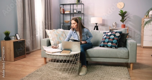 Young caucasian woman wearing wireless headset looking at laptop and making notes. Focused serious girl student watching webinar online training and talking by video conference call. Learning concept