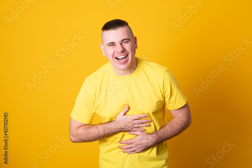 Young man in a yellow shirt holding his stomach laughing on yellow background.