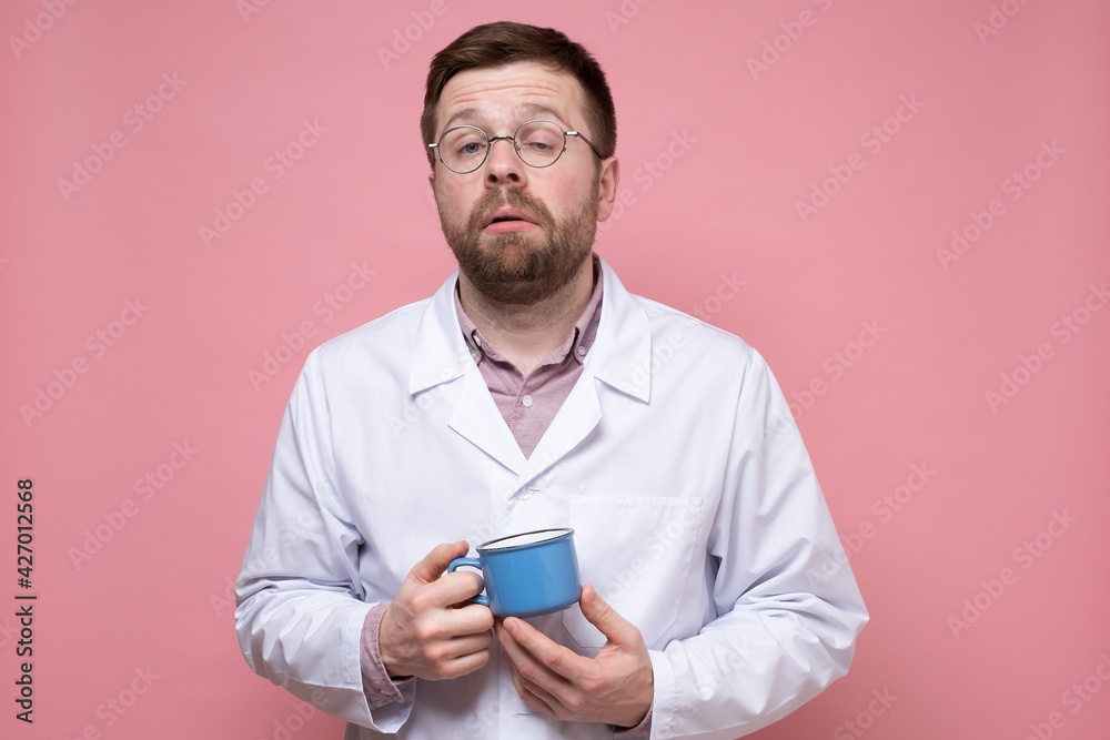 Tired Caucasian doctor holds a mug of coffee in hands, he falls asleep and tries to cheer up. Pink background. Tired Caucasian doctor holds a mug of coffee in hands, he falls asleep and tries to cheer up. Pink background.