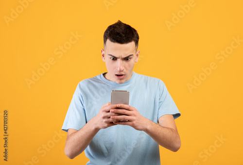 Handsome young guy staring at mobile phone in shock over orange studio background