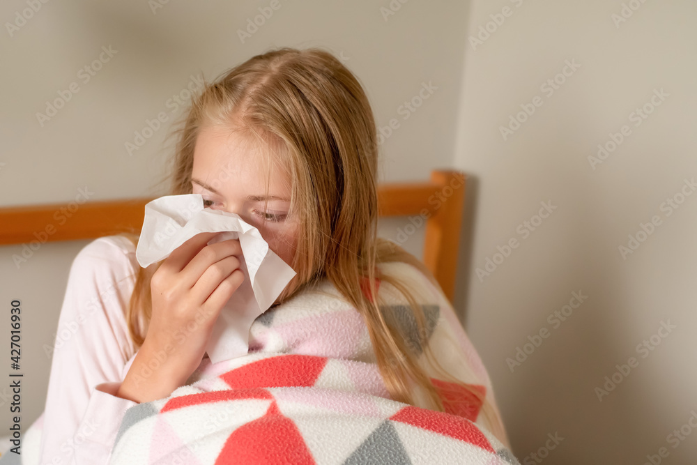 Sick teenage girl sitting on the bed covered with blanket sneezing and blowing her nose with a tissue. Sick teenage girl sitting on the bed covered with blanket sneezing and blowing her nose with a tissue.