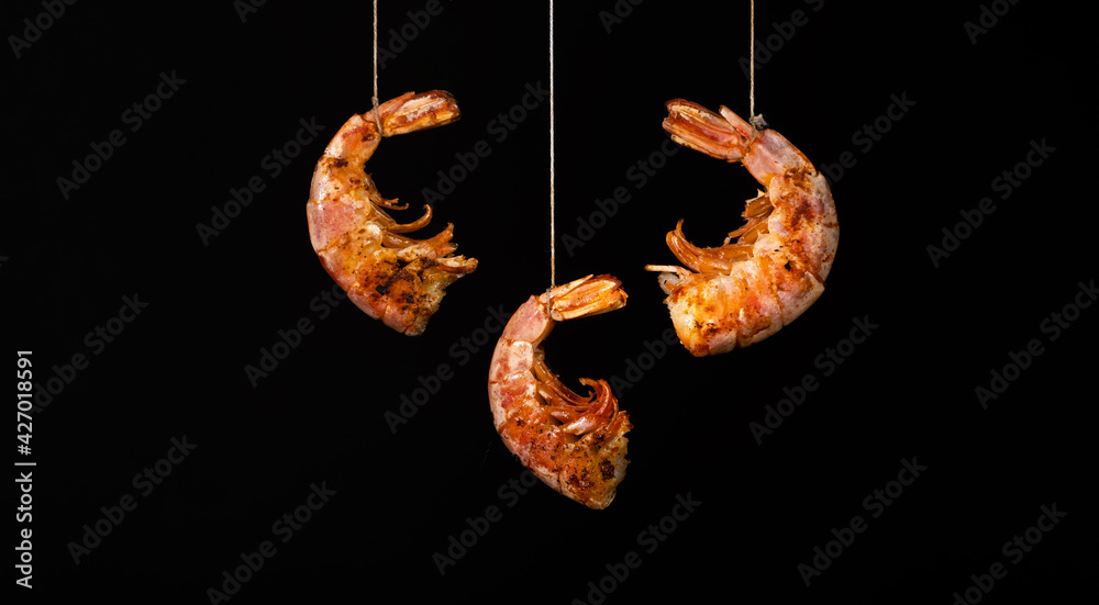 Obraz premium Fried shrimps hang on strings on a black background. Seafood appetizer, no people, horizontal orientation, copy space, close-up
