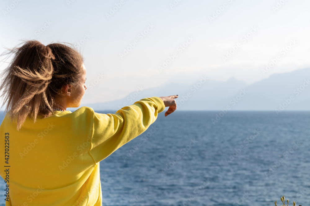 Young woman in yellow sweatshirt pointing finger at the sea.