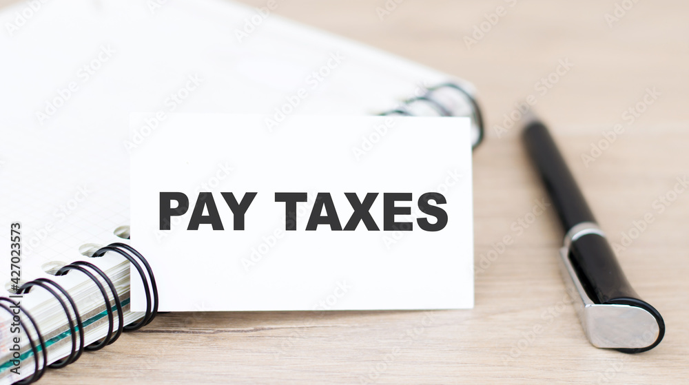 A business card with the text PAY TAXES lies on the table next to a notepad and a black pen. Business concept.