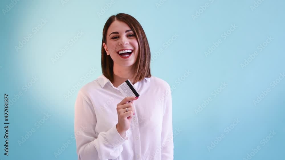 Beautiful smiling caucasian woman in white shirt shakes white credit card holding in her hand. Blue background. Studio shot. 4K resolution video. Banking and finance theme. Beautiful smiling caucasian woman in white shirt shakes white credit card holding in her hand. Blue background. Studio shot. 4K resolution video. Banking and finance theme.