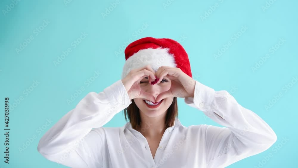 Beautiful happy caucasian woman in red Santa hat and white shirt smiles and shows heart symbol using her hands. Blue background. 4K resolution video. Positive emotion theme. Beautiful happy caucasian woman in red Santa hat and white shirt smiles and shows heart symbol using her hands. Blue background. 4K resolution video. Positive emotion theme.