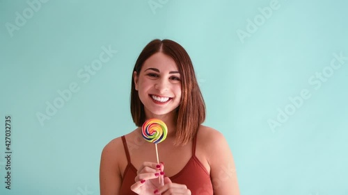 Beautiful caucasian woman with brown licks her lips while holding swirl lollipop in front of her face. 4k resolution video. Blue background. Sweet food theme.