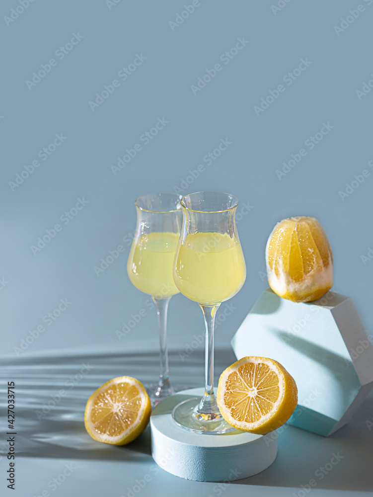 Limoncello, traditional Italian liquor on a light concrete background in the rays of the sun. Next to it is a yellow lemon, fresh citrus fruits. Composition podium.
