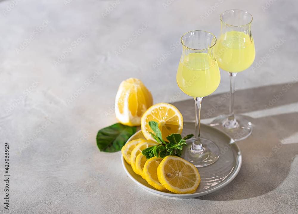 Limoncello, traditional Italian liquor on a light concrete background in the rays of the sun. Next to it is a yellow lemon, fresh citrus fruits.