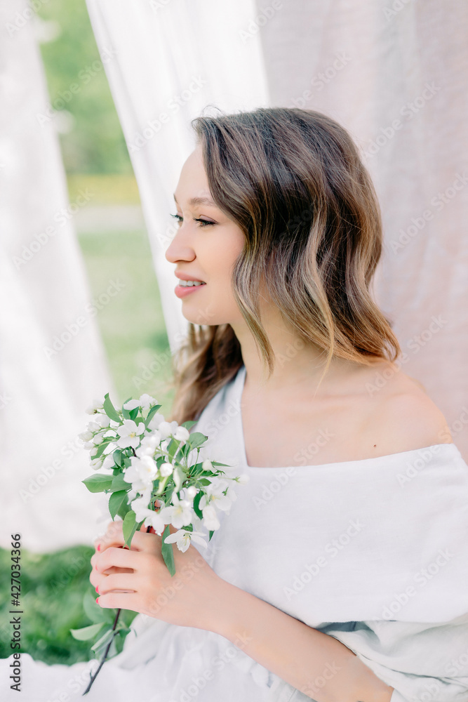 brunette woman sits on a background of linen on a rope. Spring and early summer concept. Blooming garden and a resting girl with a branch of an apple tree with flowers. brunette woman sits on a background of linen on a rope. Spring and early summer concept. Blooming garden and a resting girl with a branch of an apple tree with flowers.
