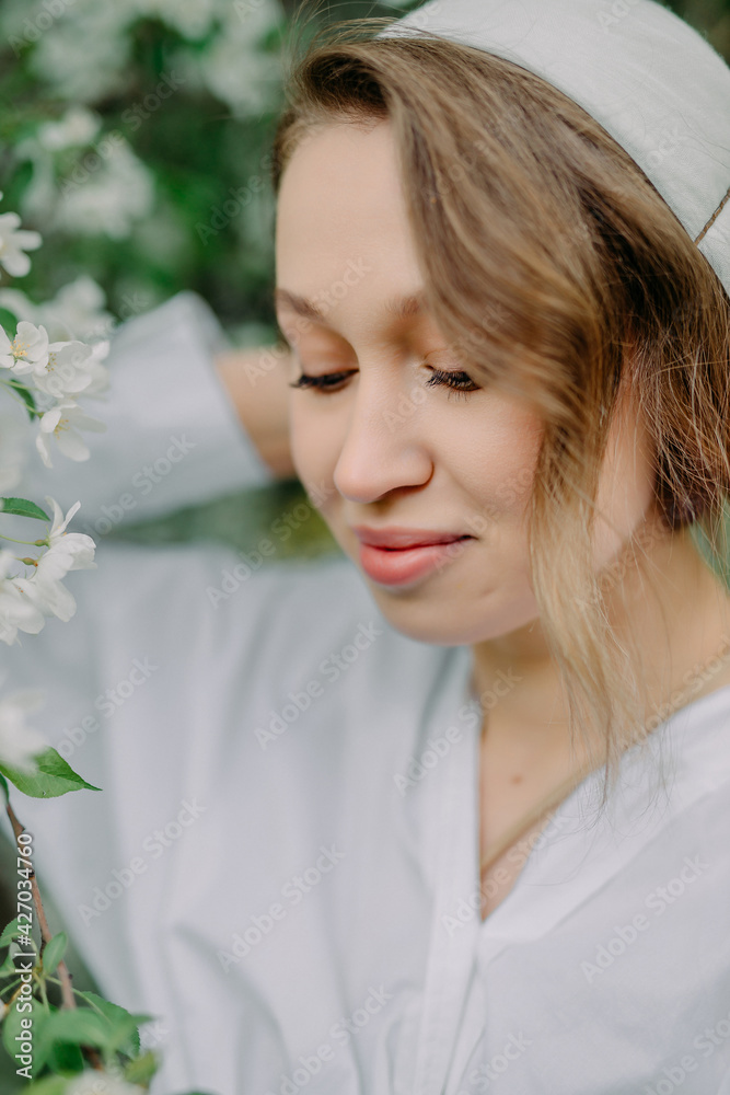 Beauty young caucasian woman in a white blouse and kerchief enjoying nature in spring apple orchard. Happy Beautiful girl walking in Garden with blooming trees. Beauty young caucasian woman in a white blouse and kerchief enjoying nature in spring apple orchard. Happy Beautiful girl walking in Garden with blooming trees.
