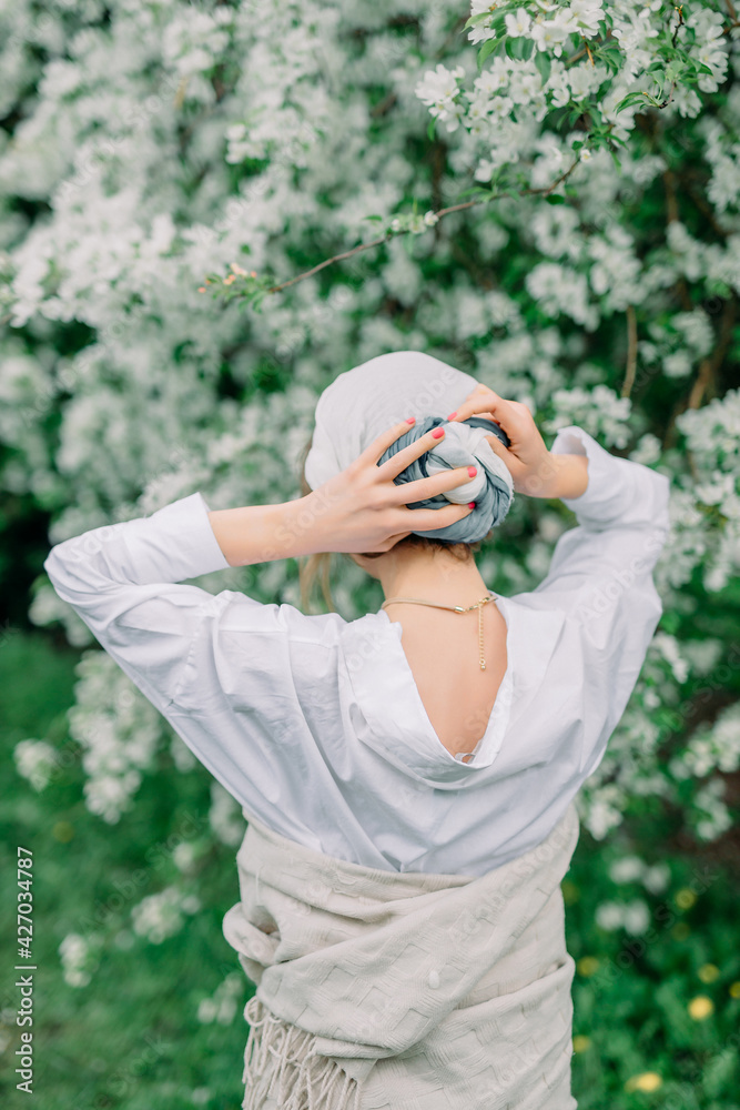 Beauty young caucasian woman in a white blouse and kerchief enjoying nature in spring apple orchard. Happy Beautiful girl walking in Garden with blooming trees. Beauty young caucasian woman in a white blouse and kerchief enjoying nature in spring apple orchard. Happy Beautiful girl walking in Garden with blooming trees.