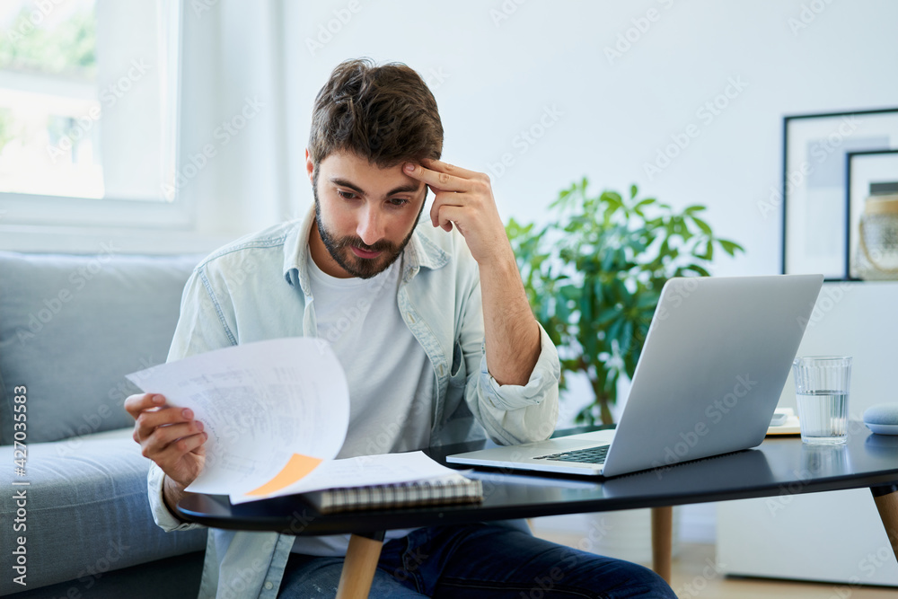 Worried young man looking on bills while at home - obrazy, fototapety, plakaty 