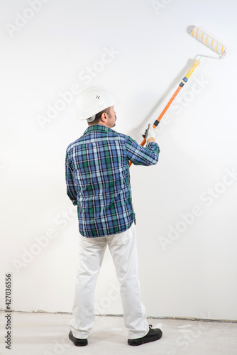 Construction worker in hard hat paints white wall with long paint roller, construction mockup,