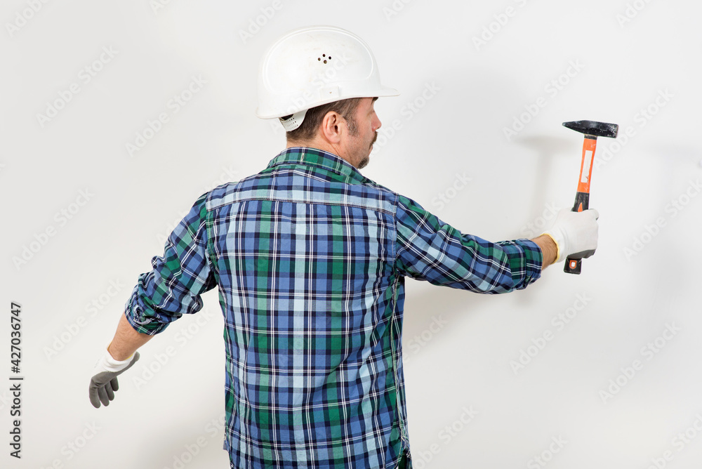 Construction worker in a protective helmet hits with a hammer against the background of a white wall Construction worker in a protective helmet hits with a hammer against the background of a white wall