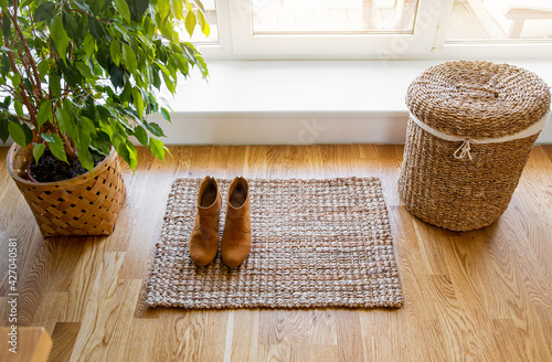 Hardwood floor with jute doormat, shoes and flower pot and seagrass laundry basket by window. Natural material objects in home concept. Home interior.  photo