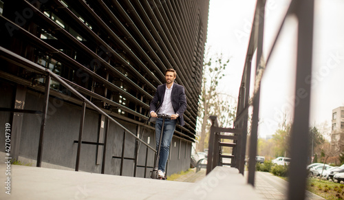 Young business man in a casual clothes riding an electric scooter by an office building on a business meeting