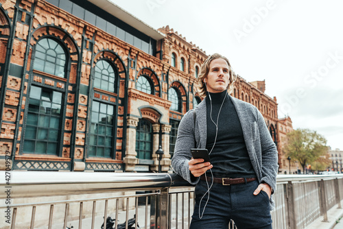 Modern businessman with smartphone in his hands on a street. Handsome man wearing earphones and speaking on mobile phone, listening music or podcast outdoors. © Rymden
