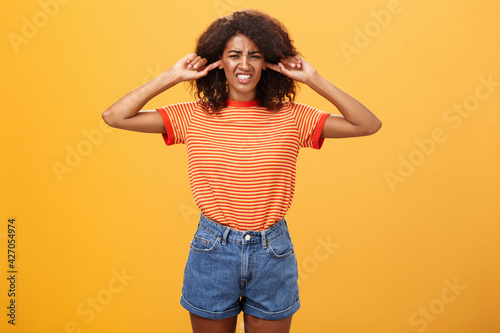 What that anoying sound. Disturbed and displeased dark-skinned girl with curly hair frowning clenching teeth from discomfort closing ears with index fingers from loud irritating sound over orange wall photo