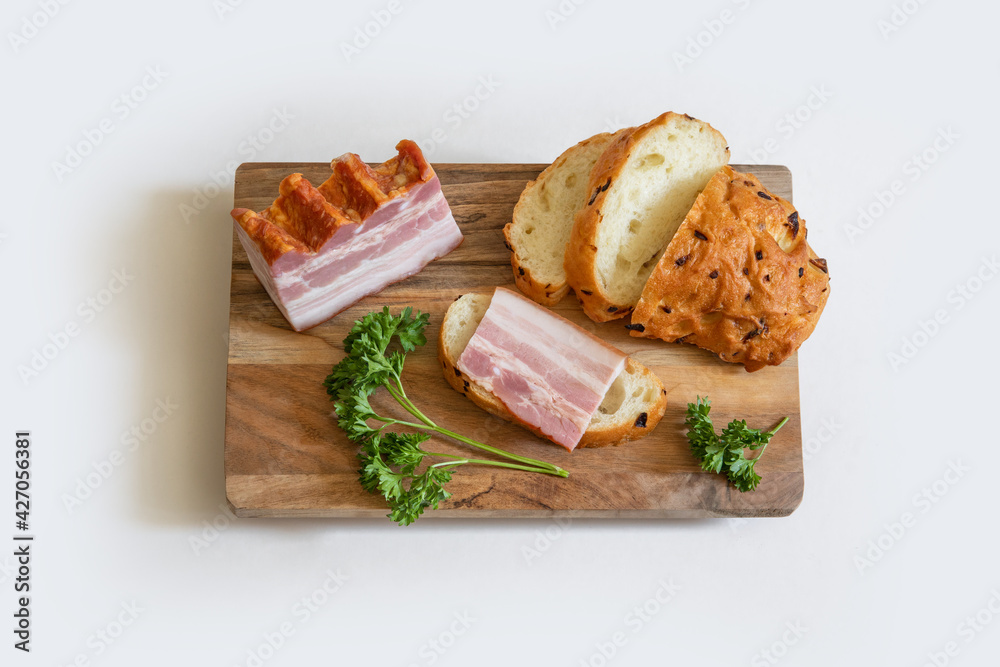 Ham slices and bread on wooden cutting board with some greens on white background.