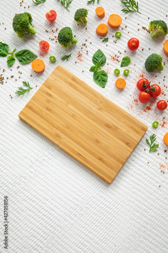 Empty wooden cutting board with vegetables and greens on white textured vintage background, copy space.
