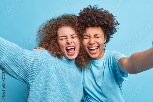 Two multicultural best female friends embrace and have fun while posing for selfie spends free time together dressed in casual clothes isolated over blue background. International friendship