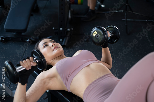 Young Asian girl in fitness gym. Asian woman working out in gym doing strength training. Fitness woman exercising indoor in fitness center. Female with dumbbells in gym. © Avirut S.